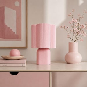 May include: A light pink table lamp with a vertically ribbed cylindrical shade and a smooth, rounded base. The lamp is positioned next to a pink vase containing small pink flowers and a stack of books topped with a pink sphere.
