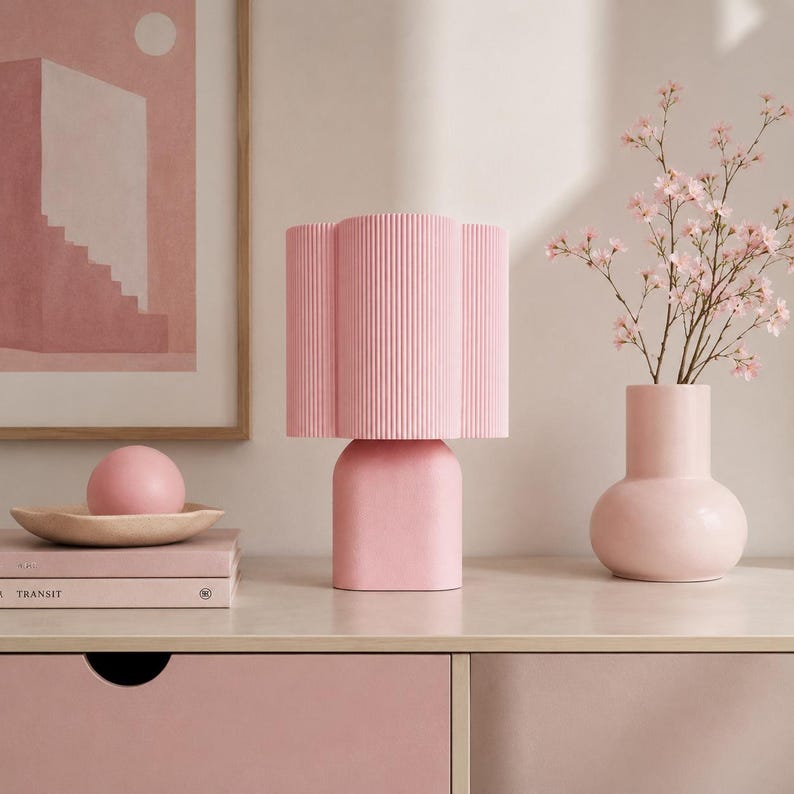 May include: A light pink table lamp with a vertically ribbed cylindrical shade and a smooth, rounded base. The lamp is positioned next to a pink vase containing small pink flowers and a stack of books topped with a pink sphere.