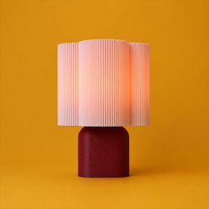 May include: A table lamp featuring a pale pink, vertically ribbed, cylindrical shade that emits a soft glow. The base of the lamp is a solid, dark red, rounded rectangular block with a textured, matte finish.