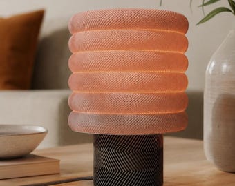 May include: A table lamp featuring a cylindrical base with a black and dark gray chevron pattern and a stacked, ribbed, translucent peach-colored shade that emits a soft, warm glow from within, resting on a flat surface.