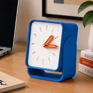 Op de afbeelding: Een vierkante analoge bureauklok met een felblauw frame en een witte wijzerplaat. De klok is voorzien van oranje uur- en minutenwijzers en eenvoudige witte streepjes als uuraanduidingen. Het ontwerp bevat een holle voet en een afgerond zijprofiel.