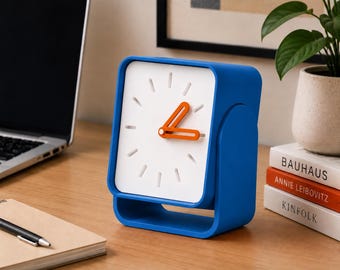 May include: A square-shaped analog desk clock with a bright blue frame and a white face. The clock features orange hour and minute hands and simple white tick marks for the hour indicators. The design includes a hollow base and a rounded side profile.