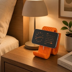 May include: A bright orange, ribbed, U-shaped phone stand holding a smartphone in landscape orientation. The phone screen displays a dark interface with an analog clock face on the left and a June calendar on the right, with the date 27 highlighted.