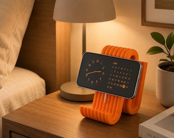 May include: A bright orange, ribbed, U-shaped phone stand holding a smartphone in landscape orientation. The phone screen displays a dark interface with an analog clock face on the left and a June calendar on the right, with the date 27 highlighted.