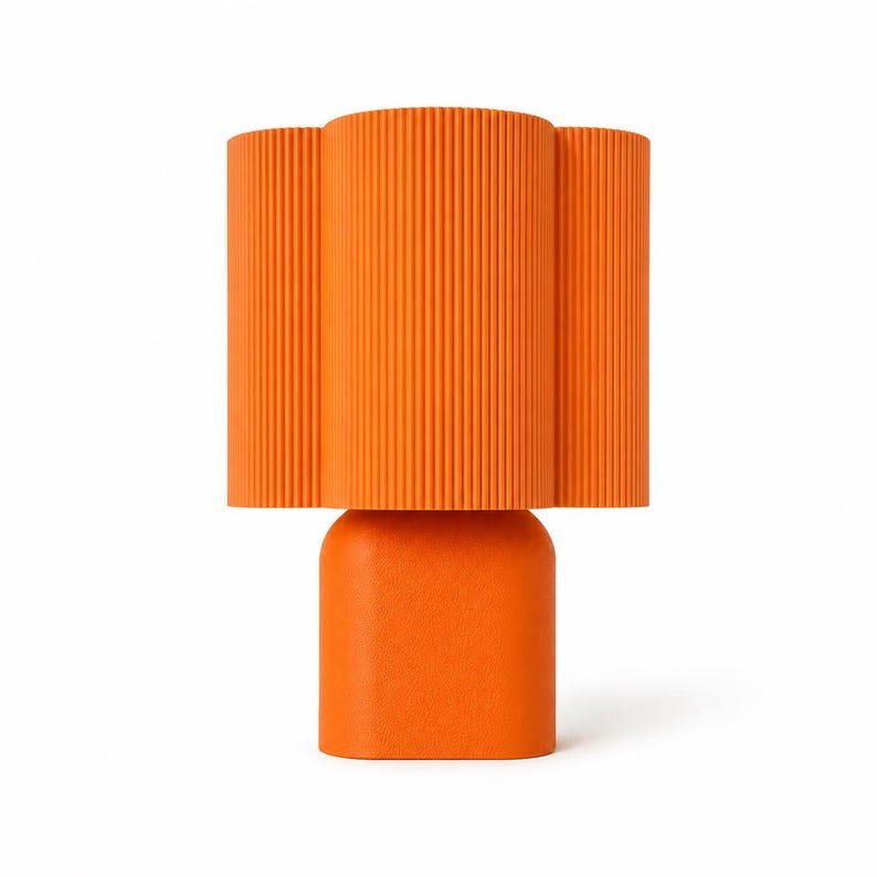 May include: A bright orange table lamp featuring a cylindrical base with a smooth matte finish and a wide, fluted lampshade with vertical ridges. The lamp has a uniform, vibrant orange color throughout its entire structure, creating a bold look.