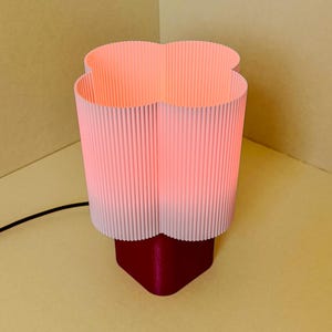 May include: A table lamp featuring a clover-shaped, light pink, vertically ribbed lampshade that emits a soft glow. The base of the lamp is a solid, dark magenta, rounded square shape. A thin black power cord extends from the base of the lamp.