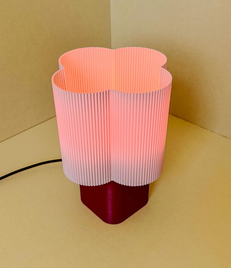 May include: A table lamp featuring a clover-shaped, light pink, vertically ribbed lampshade that emits a soft glow. The base of the lamp is a solid, dark magenta, rounded square shape. A thin black power cord extends from the base of the lamp.