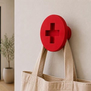 May include: A bright red wall hook shaped like a large screw head with a prominent cross-shaped slot in the centre. The circular hook is mounted to a flat surface and supports a beige canvas tote bag hanging from its lower edge.