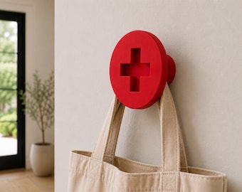 May include: A bright red wall hook shaped like a large screw head with a prominent cross-shaped slot in the center. The circular hook is mounted to a flat surface and supports a beige canvas tote bag hanging from its lower edge.
