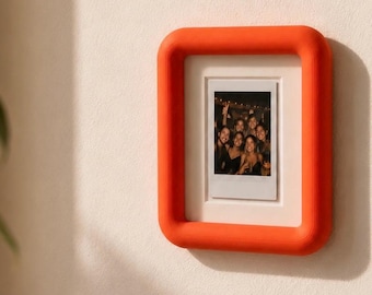 May include: A rectangular picture frame with a thick, rounded, bright orange border. The frame contains a white mat and a vertical instant film photograph showing a group of people smiling together. The frame is mounted on a plain wall.