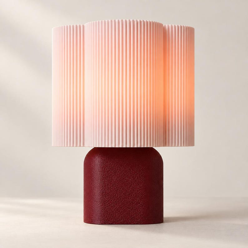 May include: A table lamp featuring a pale pink, vertically ribbed cylindrical shade with a scalloped edge. The base is a solid, deep red, rounded rectangular block with a textured, matte finish that supports the illuminated lamp shade.