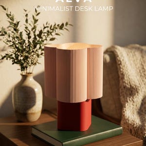 A desk lamp with a light pink, vertically ribbed, flower-shaped shade and a solid red rectangular base. The text ALVA MINIMALIST DESK LAMP by ROHËN STUDIO is visible in the upper and lower portions of the image, respectively.