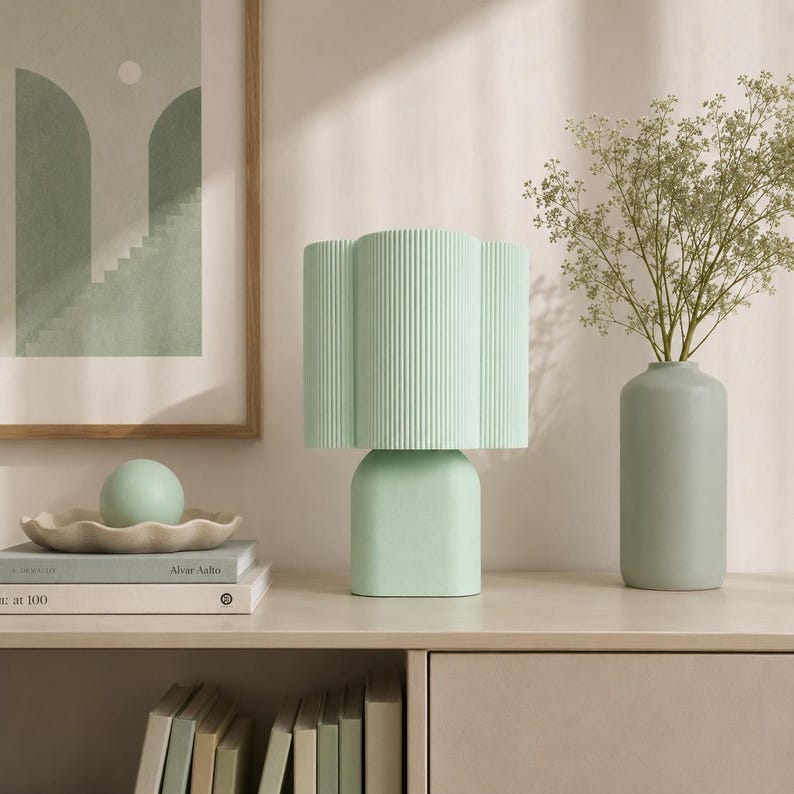 May include: A mint green table lamp with a vertically ribbed, clover-shaped shade and a smooth, rounded base. The lamp is positioned next to a tall, cylindrical mint green vase containing delicate green foliage and a small sphere.