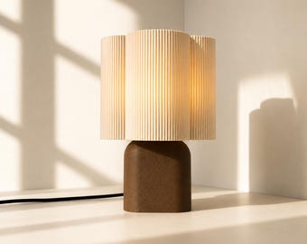 May include: A table lamp featuring a dark brown, rounded rectangular base and a cylindrical, cream-colored lampshade with a vertical ribbed texture. The lamp emits a warm, soft glow from within the shade and includes a black power cord.