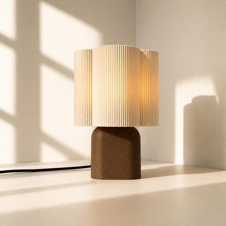 May include: A table lamp featuring a dark brown, rounded rectangular base and a cylindrical, cream-colored lampshade with a vertical ribbed texture. The lamp emits a warm, soft glow from within the shade and includes a black power cord.