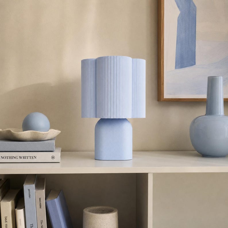 May include: A light blue table lamp with a cylindrical, vertically ribbed shade and a smooth, rounded base. The lamp is positioned on a shelf alongside a light blue sphere, a light blue vase, and several books with visible text.