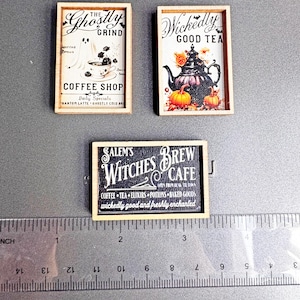 May include: Three miniature wooden signs with Halloween-themed text and illustrations. One sign reads "The Ghostly Grind Coffee Shop" with a ghost and coffee cup. Another says "Wickedly Good Tea" with a teapot and pumpkins. The third sign reads "Salem's Witches Brew Cafe".