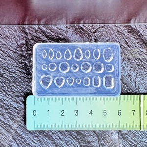 May include: A clear silicone mold with various shapes, including teardrops, ovals, and hearts. The mold is rectangular and sits next to a ruler, with measurements in inches. Ideal for crafting jewelry or decorative elements.