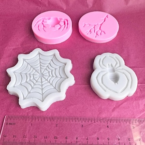 May include: Four silicone molds in pink and white, featuring Halloween-themed designs. The molds include a spider, spiderweb, and two circular molds with a crab and a tree branch design. A ruler is placed below the molds.