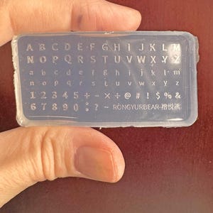 May include: A clear, rectangular silicone mold with embossed letters, numbers, and symbols. The mold includes uppercase and lowercase alphabets, numbers 1-0, and various symbols. The text "RONGYURBEAR-榕悦熊" is also visible.