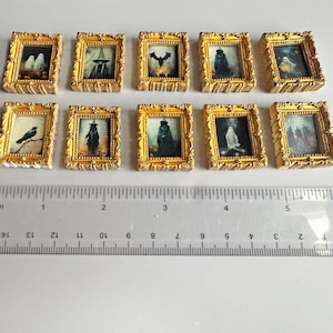 Puede incluir: Diez pinturas en miniatura con marcos dorados con varias imágenes espeluznantes, incluyendo una bruja, un murciélago y un fantasma. Una regla con medidas en centímetros se encuentra debajo.