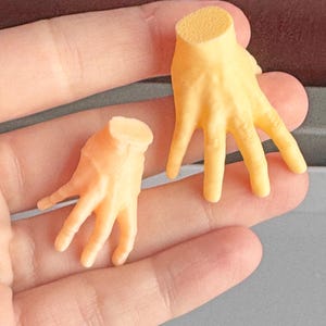 May include: Two small, light orange, 3D-printed hands. One hand is open, and the other is closed. The hands are cut off at the wrist. The hands are resting on a light orange surface.