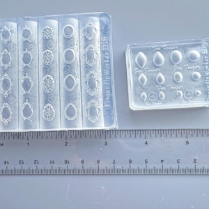May include: Two clear silicone molds with various decorative shapes, including oval frames and small geometric designs. A ruler with inch measurements is placed below the molds, showing a length of approximately 6 inches.