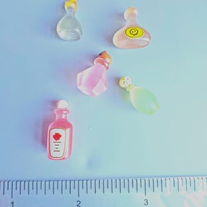 Op de afbeelding: Een verzameling kleine, kleurrijke toverdrankflesjes. De flesjes hebben verschillende vormen en kleuren, waaronder transparant, roze en groen. Sommige hebben gele stoppen en één heeft een etiket met een rode roos. Een liniaal staat op de voorgrond.