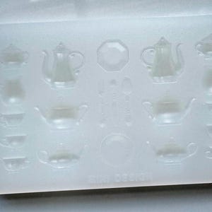 Puede incluir: Un molde de silicona transparente y rectangular con juegos de té en miniatura. El molde incluye teteras, tazas de té, azucareros y platos. Las palabras "MINI DESIGN" están impresas en la parte inferior.