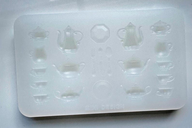 May include: A clear, rectangular silicone mold featuring miniature tea sets. The mold includes teapots, teacups, sugar bowls, and serving plates. The words "MINI DESIGN" are printed at the bottom of the mold.
