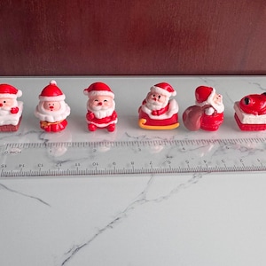 Puede incluir: Colección de seis figuras en miniatura de Papá Noel. Cada figura viste un traje rojo con barba y gorro blancos. Una regla con medidas en pulgadas se coloca debajo.
