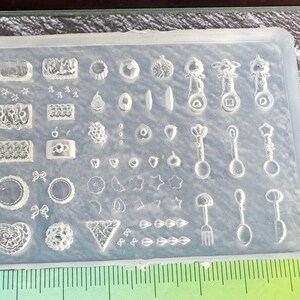 May include: A clear silicone mould filled with various shapes for crafting. The mould contains miniature designs including stars, bows, spoons, and geometric shapes. The mould is rectangular and has a grid of different shapes.