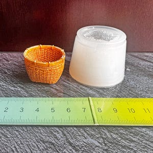 May include: A miniature orange woven basket next to a translucent silicone mold. A green ruler is positioned in the foreground, showing the basket has a diameter of approximately 1.5 inches and the mold has a diameter of approximately 2 inches.