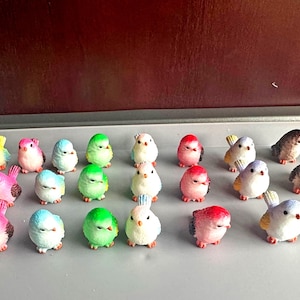 May include: A selection of small, colourful bird figurines in shades of yellow, pink, blue, green, red, and brown. The birds are displayed on a grey surface, creating a charming and decorative arrangement.