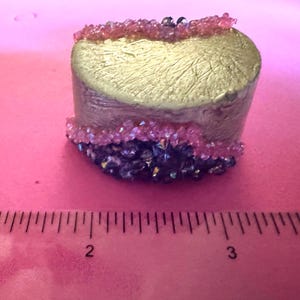 May include: A small, cylindrical decorative object with a gold-coloured top and a textured surface. The sides are a muted metallic colour, and it is adorned with pink and dark purple crystals. The object is approximately 2.5 cm in diameter.