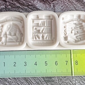 Könnte beinhalten: Eine weiße Silikonform mit drei rechteckigen Vertiefungen, die geprägte Motive einer Leseecke, eines Bücherregals und eines Bücherstapels zeigen, platziert über einem grünen Lineal mit einer Länge von etwa 10 Zentimetern.