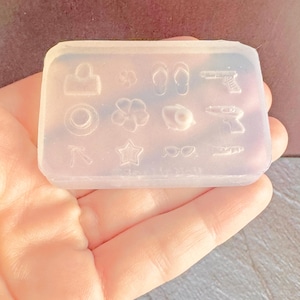 以下が含まれることがあります： 帽子、ビーチサンダル、銃、星、サングラスなどのミニチュア形状が特徴の透明なシリコンモールド。長方形のモールドは手で持たれており、背景はぼやけています。
