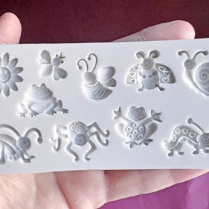 Può includere: Uno stampo in silicone bianco con disegni in rilievo di vari insetti e un fiore. Lo stampo include un'ape, una farfalla, una coccinella, una lumaca, una rana, un ragno e un bruco. Lo stampo è tenuto in una mano.