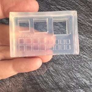 Puede incluir: Un molde de silicona transparente y rectangular con un diseño de ventana y rejilla. El molde tiene tres aberturas rectangulares en forma de ventana en la parte superior y una rejilla de pequeños cuadrados en la parte inferior. El texto "9910!" es visible.