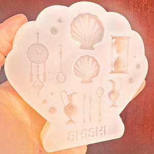 May include: A seashell-shaped silicone mold with various detailed designs, including seashells, an hourglass, a clock, and serving utensils. The word "SHISHI" is at the bottom. The mold is held in a hand.