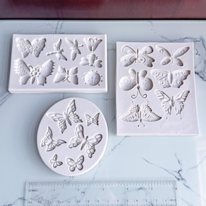 Puede incluir: Tres moldes de silicona blancos con diseños de mariposas. Un molde rectangular presenta varias formas de mariposas e insectos. Otro molde rectangular tiene diferentes diseños de mariposas. Un tercer molde ovalado muestra varias formas de mariposas. Se incluye una regla.