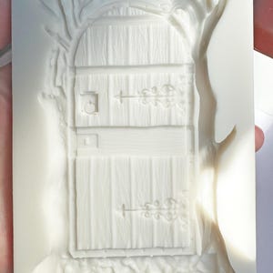 May include: A white silicone mold featuring a detailed fairy door design. The door has a wooden plank texture, decorative hinges, and a small handle. A tree and foliage frame the door, creating a whimsical, fantasy-themed item.