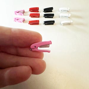 May include: A pink miniature stapler is held in a hand, with several other staplers in red, black, and white arranged in the background. The staplers are small, likely for a dollhouse or craft project.