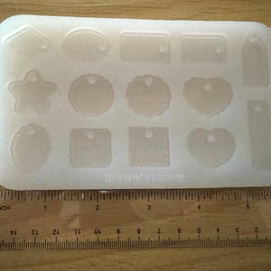 May include: A clear, rectangular silicone mold with various shapes, including tags, hearts, and stars. The mold is placed on a wooden surface with a ruler showing inches. The text "moon/vroom" is visible on the mold.