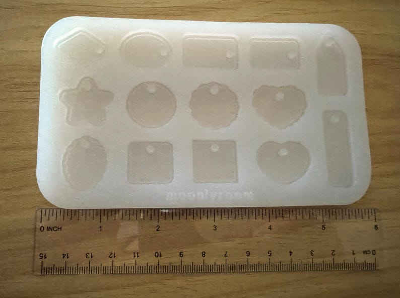 May include: A clear, rectangular silicone mold with various shapes, including tags, hearts, and stars. The mold is placed on a wooden surface with a ruler showing inches. The text "moon/vroom" is visible on the mold.