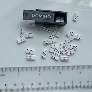 May include: A black box labeled "DOMINO" contains white dominoes with black dots. Several dominoes are scattered around the box. A ruler shows measurements in inches, with the dominoes appearing to be approximately 1 inch in length.