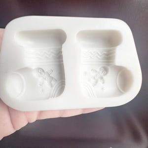 Puede incluir: Un molde de silicona blanco con dos formas de calcetines navideños. Cada calcetín tiene detalles en relieve, incluyendo un patrón en zigzag en la parte superior, copos de nieve y un lazo. El molde es sostenido por una mano.