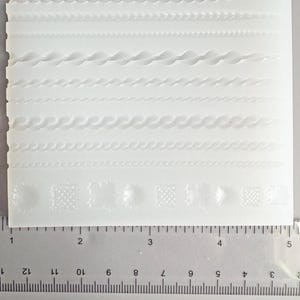 May include: A white silicone mold featuring various decorative border designs. The mold includes patterns like waves, braids, and geometric shapes. A ruler is placed next to the mold, with measurements in inches.