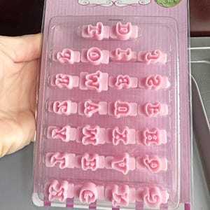 Può includere: Mini taglia biscotti a forma di alfabeto maiuscolo rosa in una confezione di plastica trasparente. La confezione ha uno sfondo a righe rosa e bianche. I taglia biscotti sono disposti in file e sono in plastica rosa. Con il testo "Mini Uppercase Alphabet Cutters".