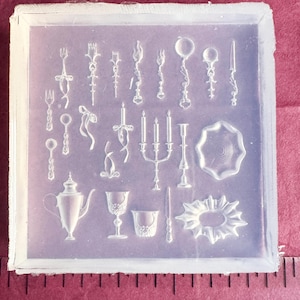 May include: A clear silicone mold with various miniature kitchen and dining items. The mold includes forks, spoons, candles, a teapot, glasses, and a decorative plate. The items are arranged in a grid pattern within the square mold.